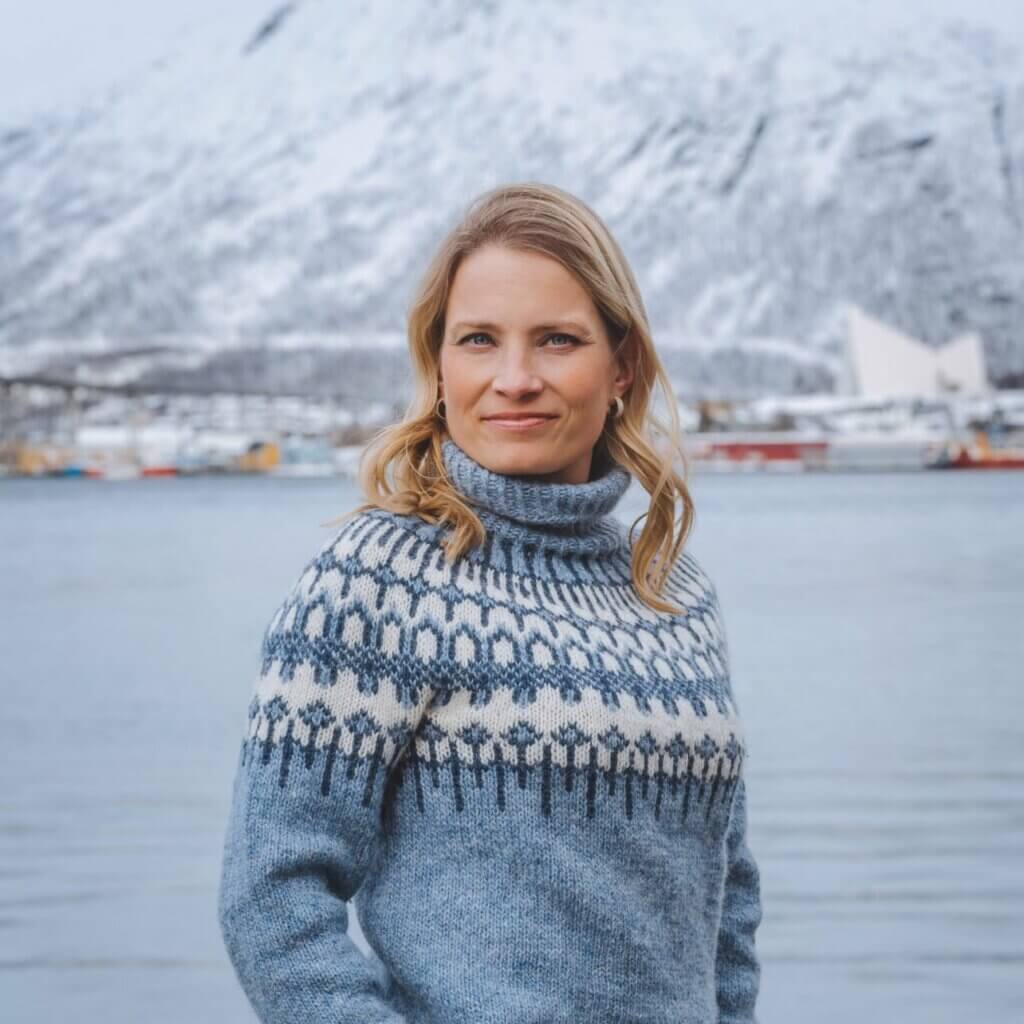 Image shows a blonde woman in a woolen sweater outdoors. It is outdoors and the scenery shows parts of Tromsø city.