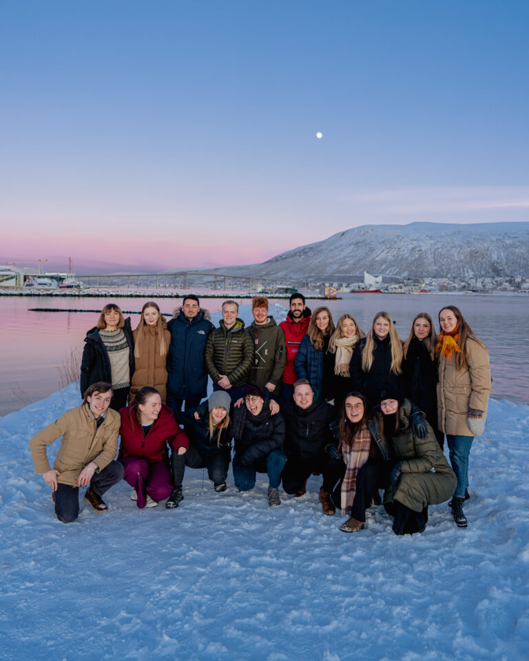 Arctic Frontiers - A group of people from the Arctic Frontiers - Young Student Forum pose on snowy ground by a lake, with mountains and a sunset sky in the background.