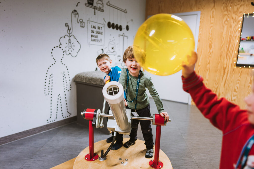Et barn holder en gul ballong foran to andre barn som smiler. De to barna styrer ballongen med luft fra en 