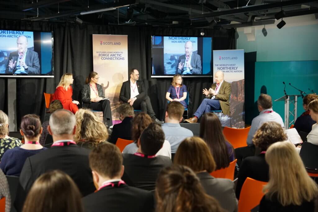 Image shows five people on the stage and several people in the audience listening.