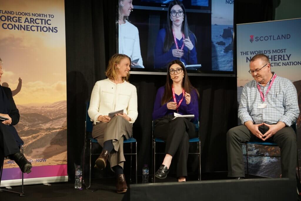 Image shows three people on the stage engaging in a panel discussion. 