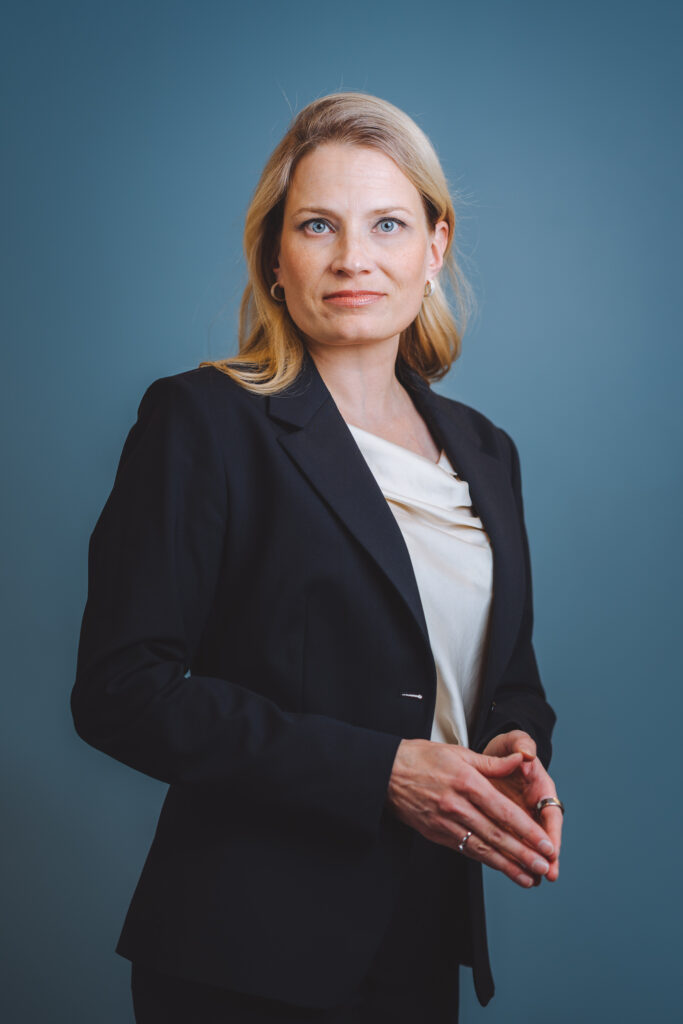 Image shows blonde woman in black suit, with a serious face.