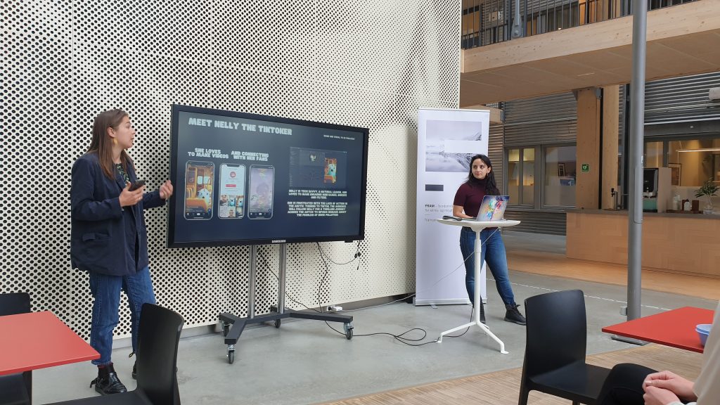 Two women stand either side of a TV screen with various pictures of the app they have developed.