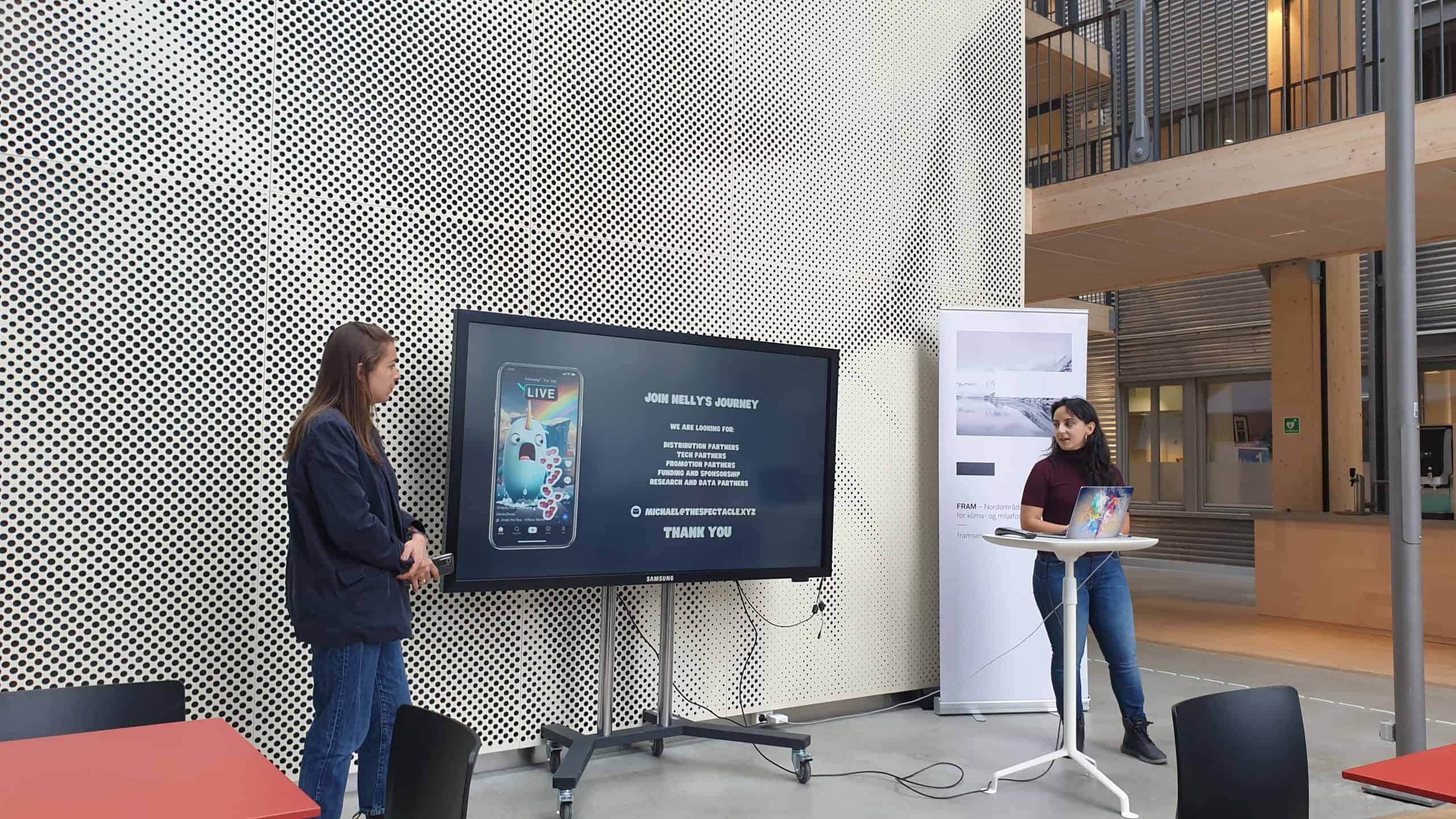Two young women stand either side of a TV screen which shows a photo of a mobile phone with an app showing Nelly the Nahwahl.