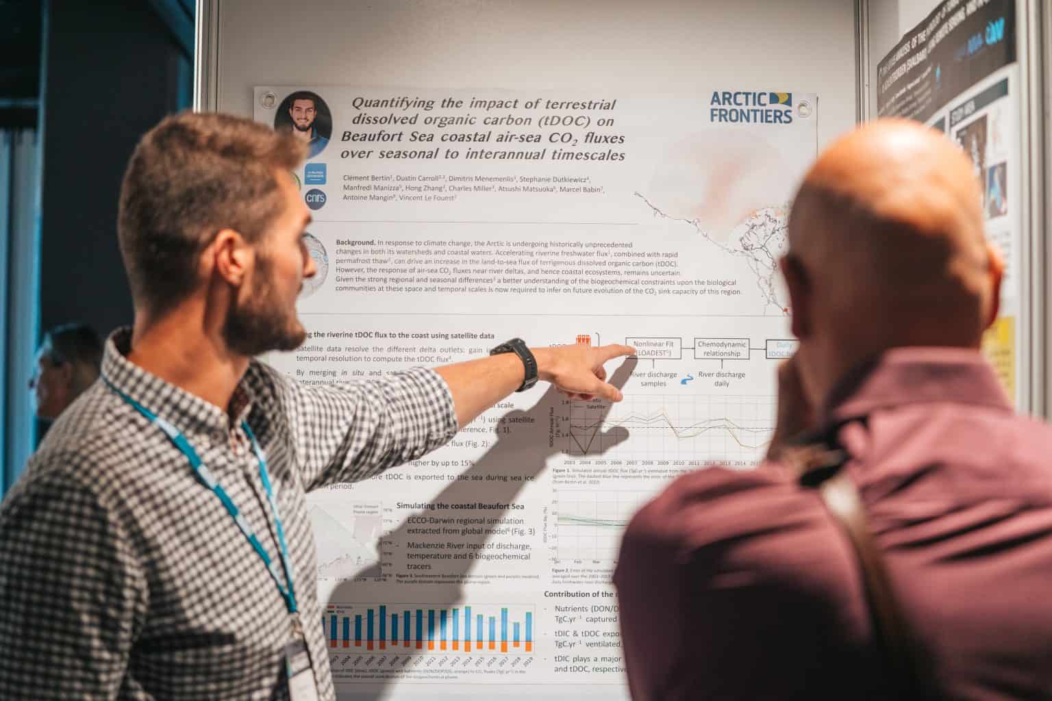 Two men stand beside a scientific poster. The photo is shot from behind them. The man on the left points at something on the poster.