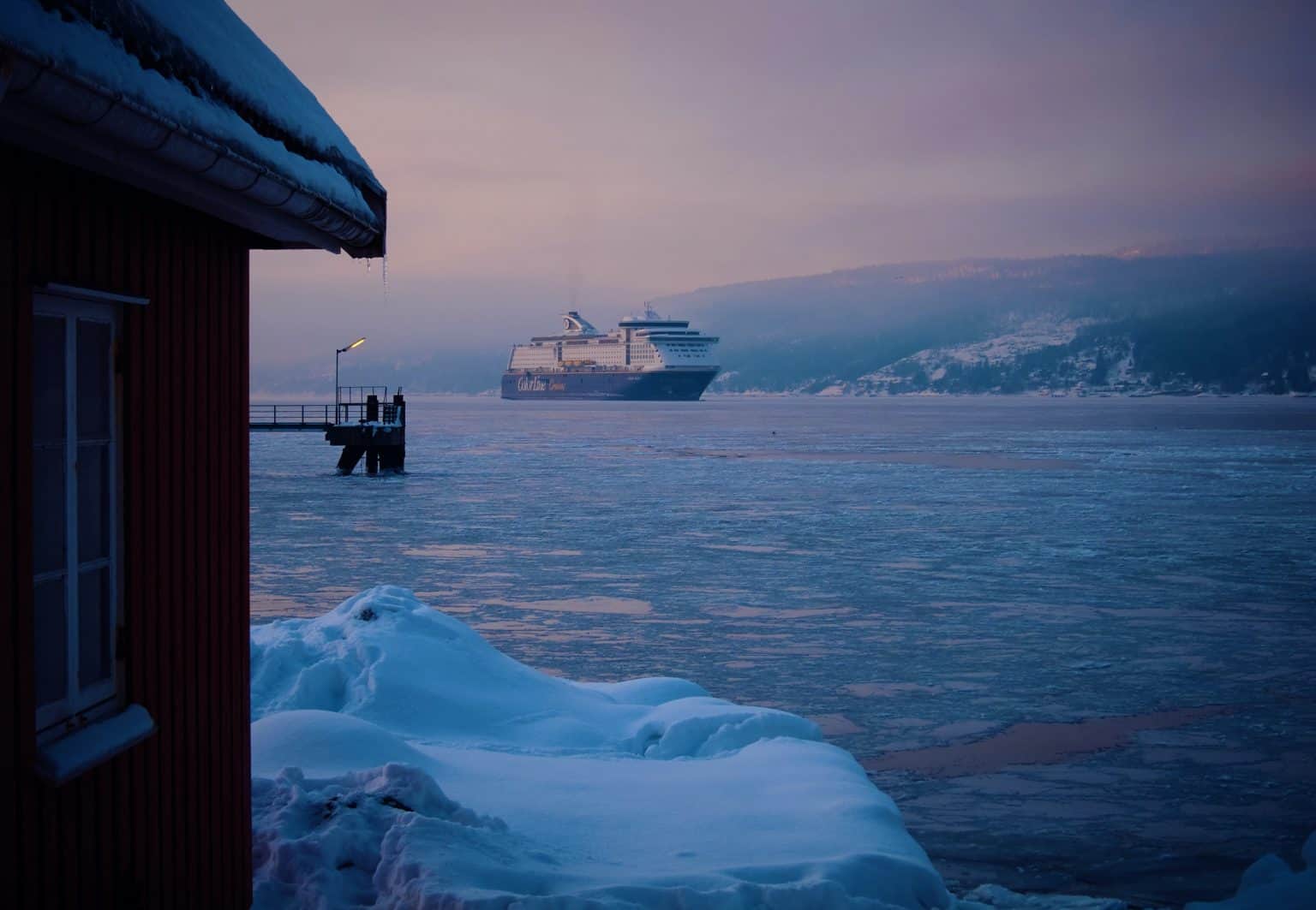 A cruise ship in the background, floating in a fjord. In the foreground, a hut with snow on the roof and sea ice in the fjord.