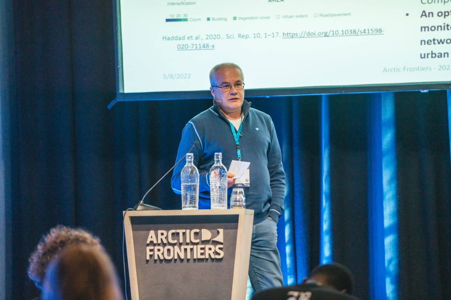 A male scientist stands on stage behind a podium with the Arctic Frontiers logo. He is presenting his work to the audience.