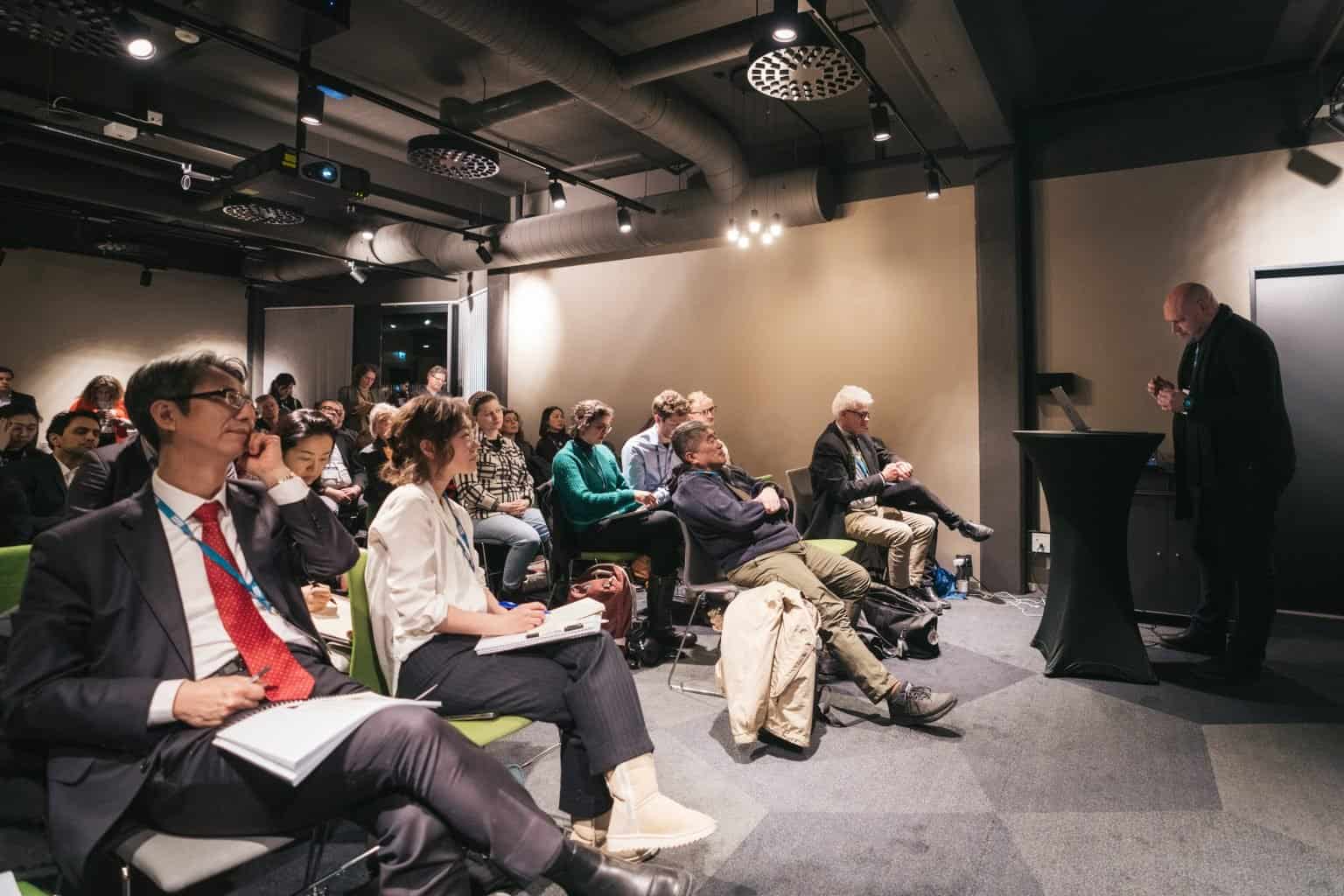 Scientists gather in a room to listen to another talk about their work. Photo taken from the front of the room where you can see an audience of around 25 people.