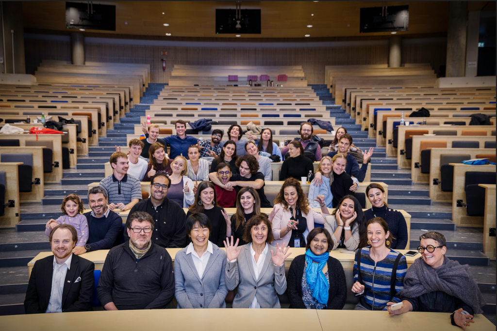 A group of students and professors sit in a lecture hall smiling and pulling silly faces.