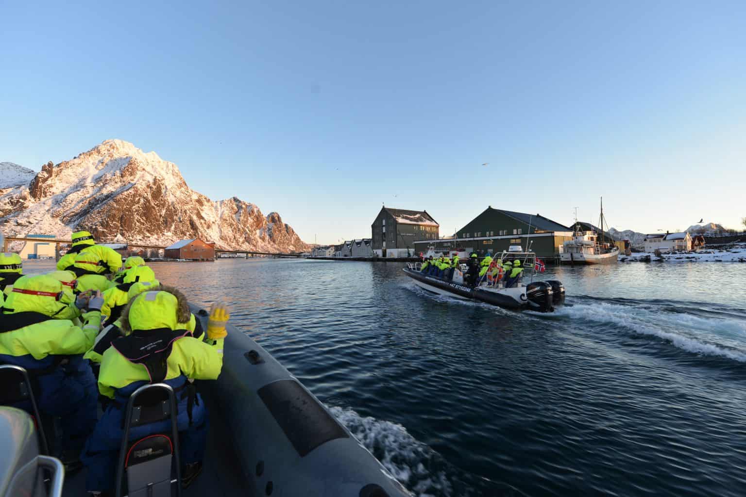 Arctic Frontiers - People in yellow jackets, part of Arctic Frontiers - Emerging Leaders - 2022 and older, ride inflatable boats on calm water near snowy mountains and buildings.