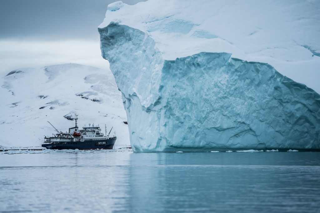 Image shows a ship in front of a massive iceberg in Greenland.