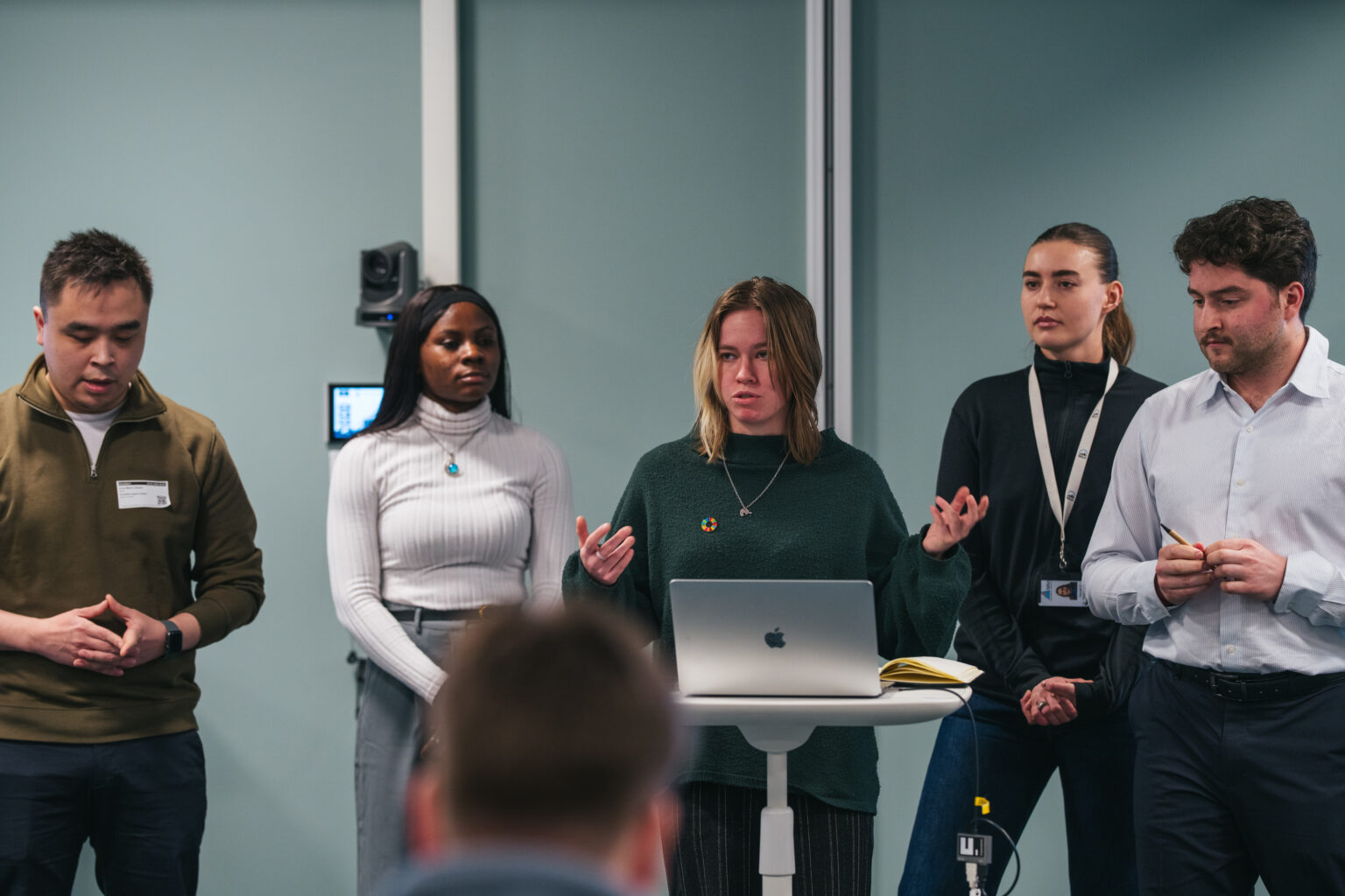 Arctic Frontiers - Five people stand at the front of a room during a forum; one student speaks at a lectern with a laptop while the others listen attentively.