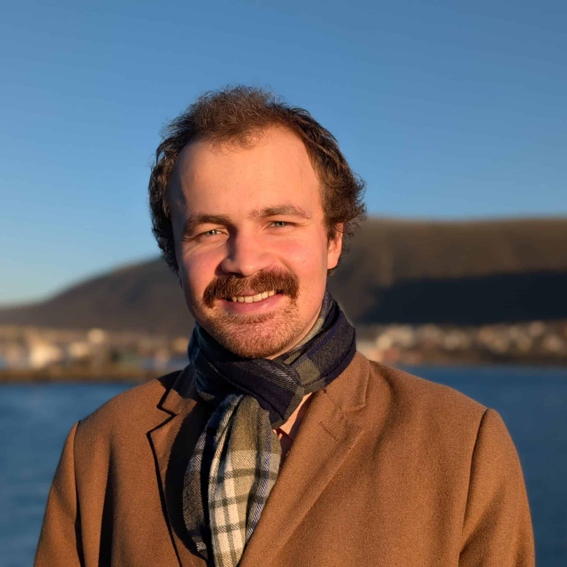 Arctic Frontiers - Thomas Helland-Hansen with a moustache, wearing a brown coat and a scarf outdoors by the water.