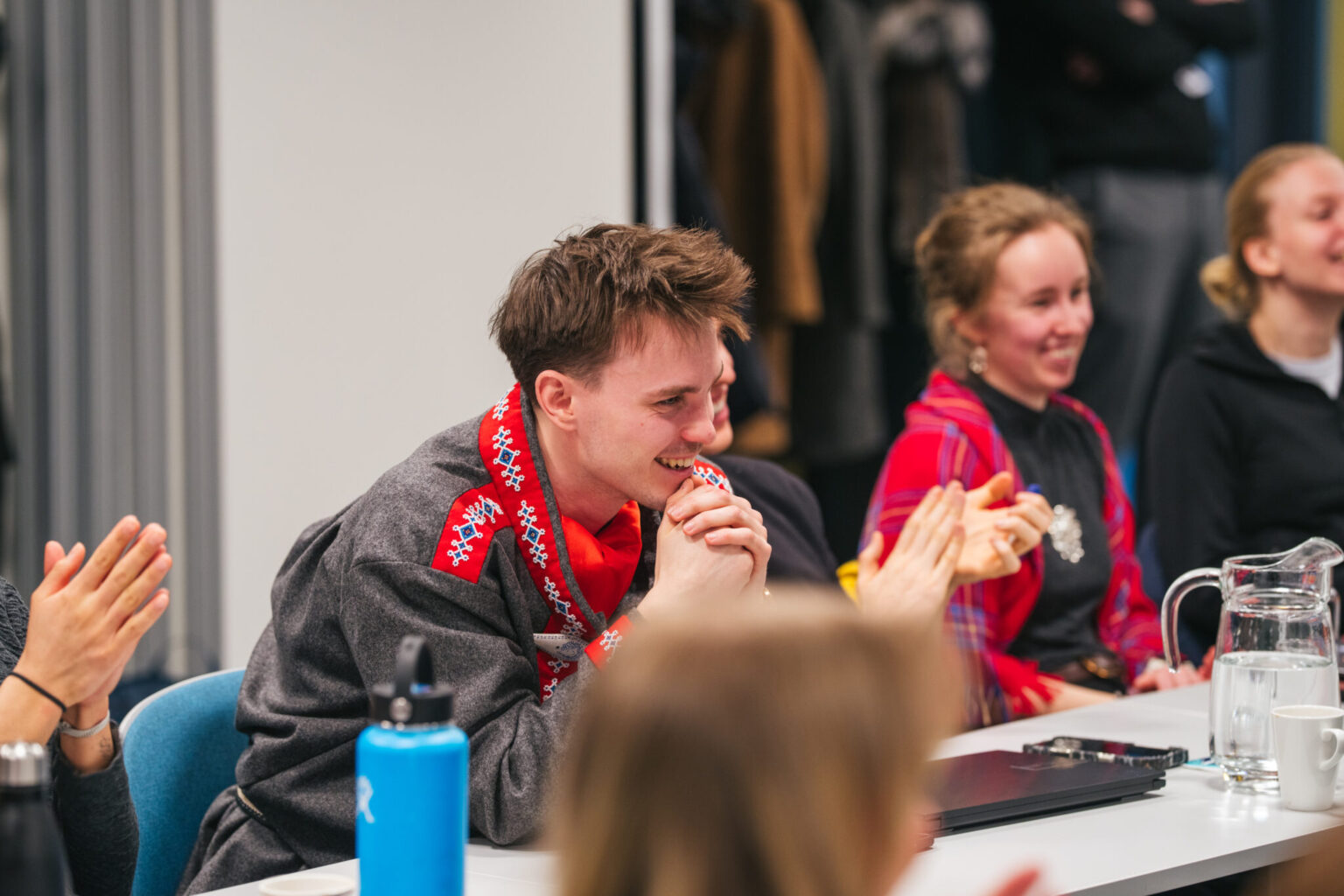 Arctic Frontiers - Smiling students sit at a table, clapping and listening, with water bottles and a jug nearby.