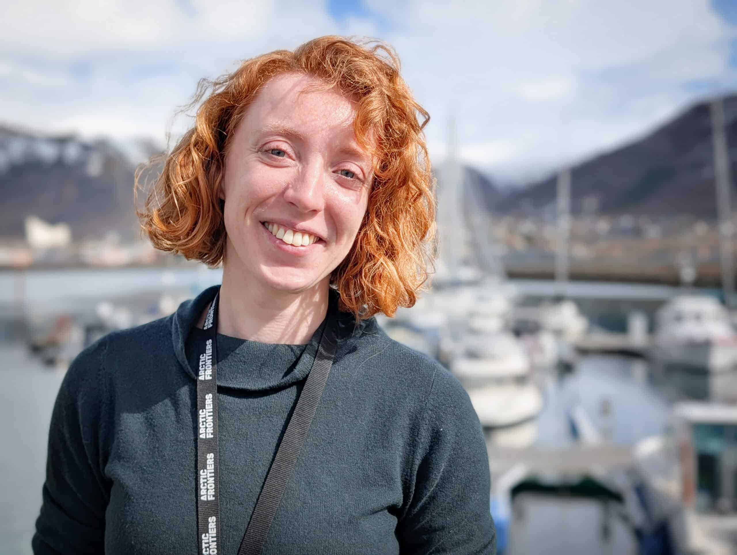 Arctic Frontiers - Smiling person with curly red hair stands by a harbour with boats and mountains in the background.