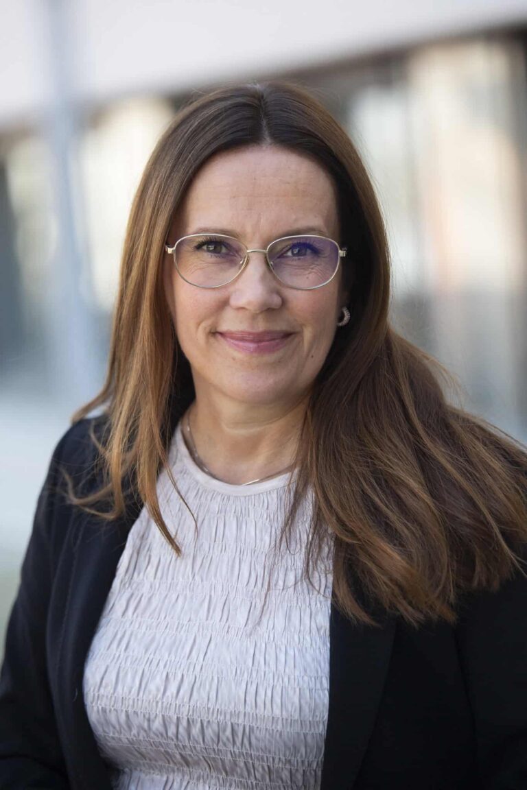 Arctic Frontiers - Woman with long brown hair, wearing glasses, a light top, and a dark blazer, smiling outdoors.