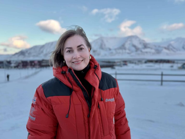 Arctic Frontiers - Woman in a red winter jacket standing in a snowy landscape with mountains in the background.