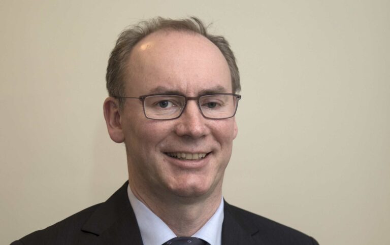 Arctic Frontiers - Man with glasses in a suit and tie smiling in front of a plain light-coloured background.