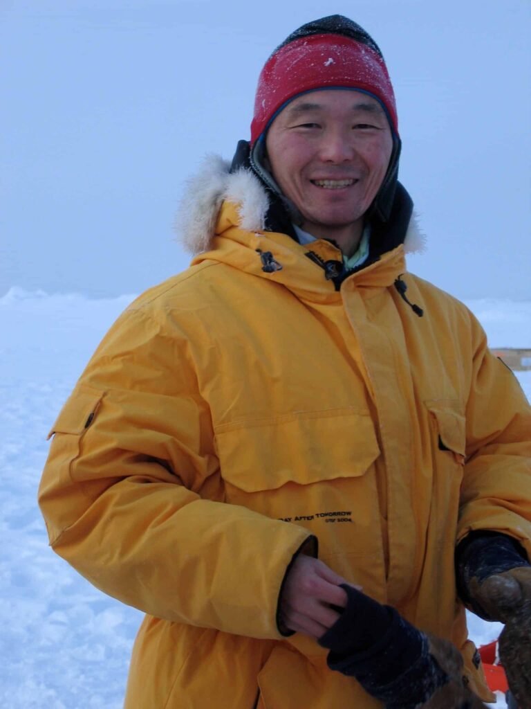 Arctic Frontiers - Smiling person in a yellow anorak and red hat standing outside on a snowy, icy landscape.