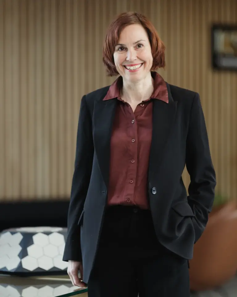 Arctic Frontiers - Woman with short brown hair in a black suit and maroon shirt smiles, standing indoors near a table.