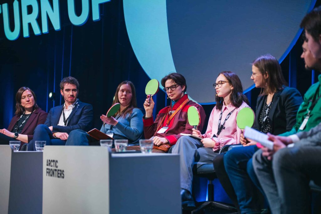 Arctic Frontiers - Seven panellists sit on stage, some holding green paddles, engaged in a discussion at a conference.