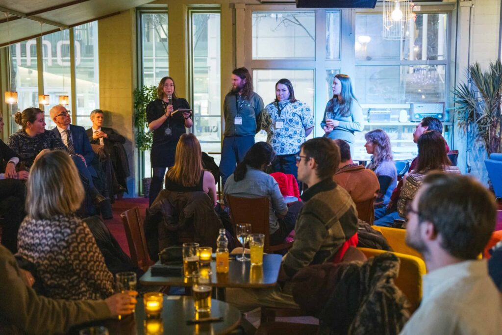 Arctic Frontiers - Four people stand speaking to a seated audience in a cosy, well-lit room with drinks on tables.