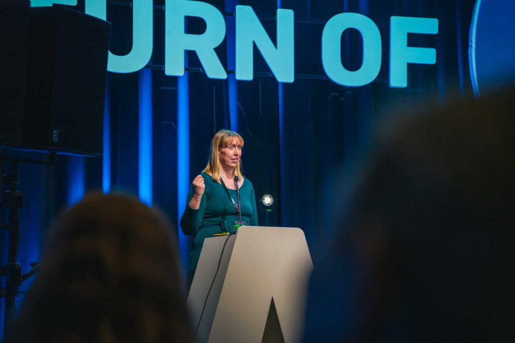 Arctic Frontiers - Woman speaking at lectern on stage with large “URN OF” letters illuminated behind her.
