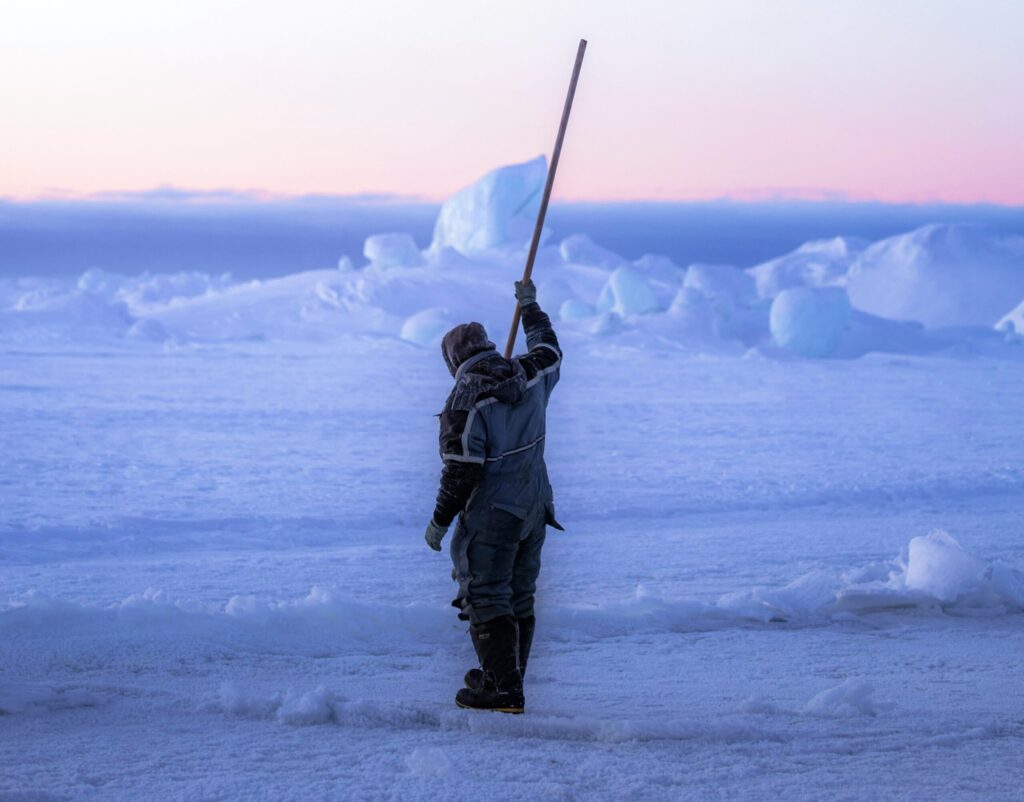 Arctic Frontiers - A person in winter gear stands on snow, holding a long pole upwards with icebergs in the background.