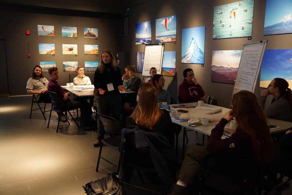 Arctic Frontiers - A group of people sit around tables, listening to a woman standing and speaking in a room with holiday photos.