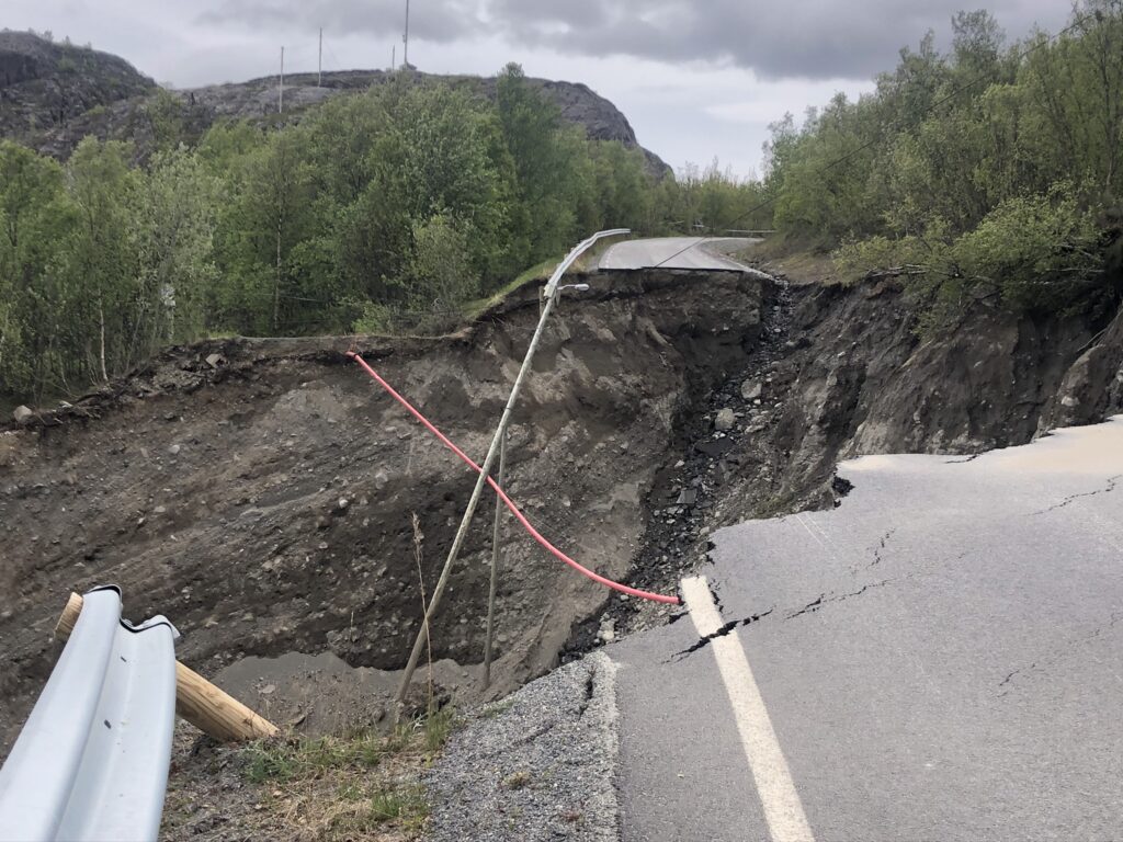 Arctic Frontiers - A road is severely eroded, creating a large gap, with broken crash barriers and exposed earth.