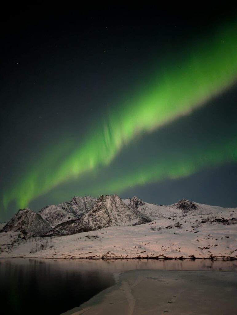 Arctic Frontiers - Green northern lights glow above snowy mountains and a calm lake under a dark night sky.