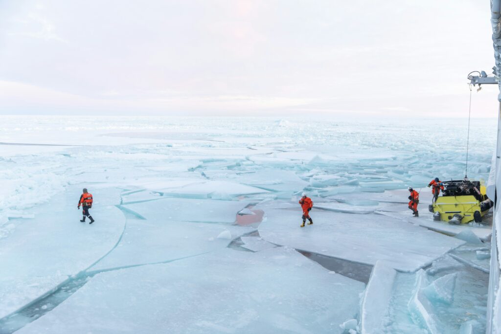 Arctic Frontiers - Four people in orange suits walk on cracked ice near a yellow vehicle and a ship in a frozen landscape.