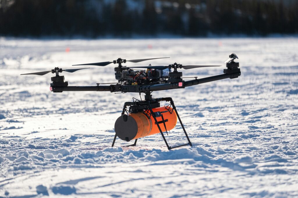 Arctic Frontiers - A large drone with an orange container attached, standing on snow in a sunny outdoor setting.