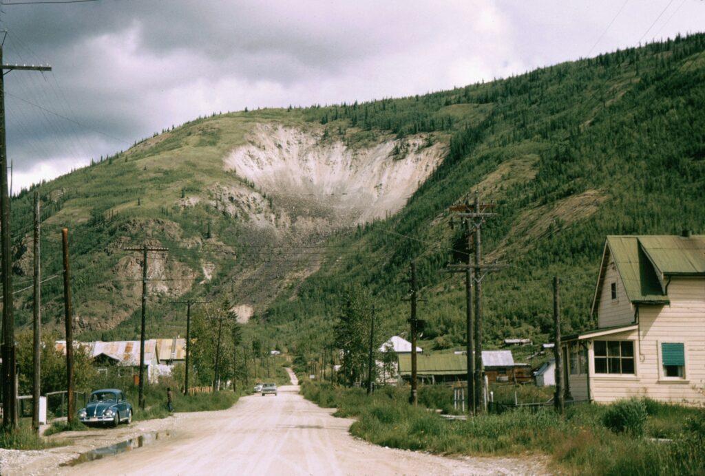 Arctic Frontiers - A dirt road leads towards a green hill, with houses, power lines, and cars along the roadside.