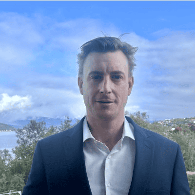 Arctic Frontiers - Man in a dark suit and white shirt standing outside with trees, water, and mountains in the background.
