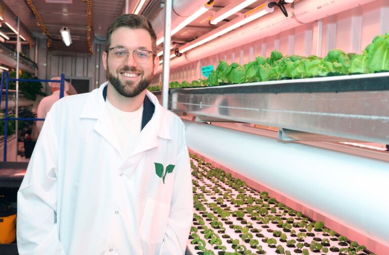 Arctic Frontiers - A smiling man in a lab coat stands in a North Halifax vertical farm with shelves of leafy greens.