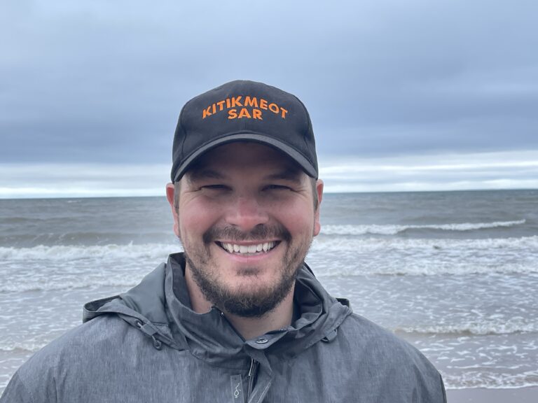 Arctic Frontiers - A smiling man wearing a “KITIKMEOT SAR” baseball cap on an Arctic beach, embodying Northern resilience.