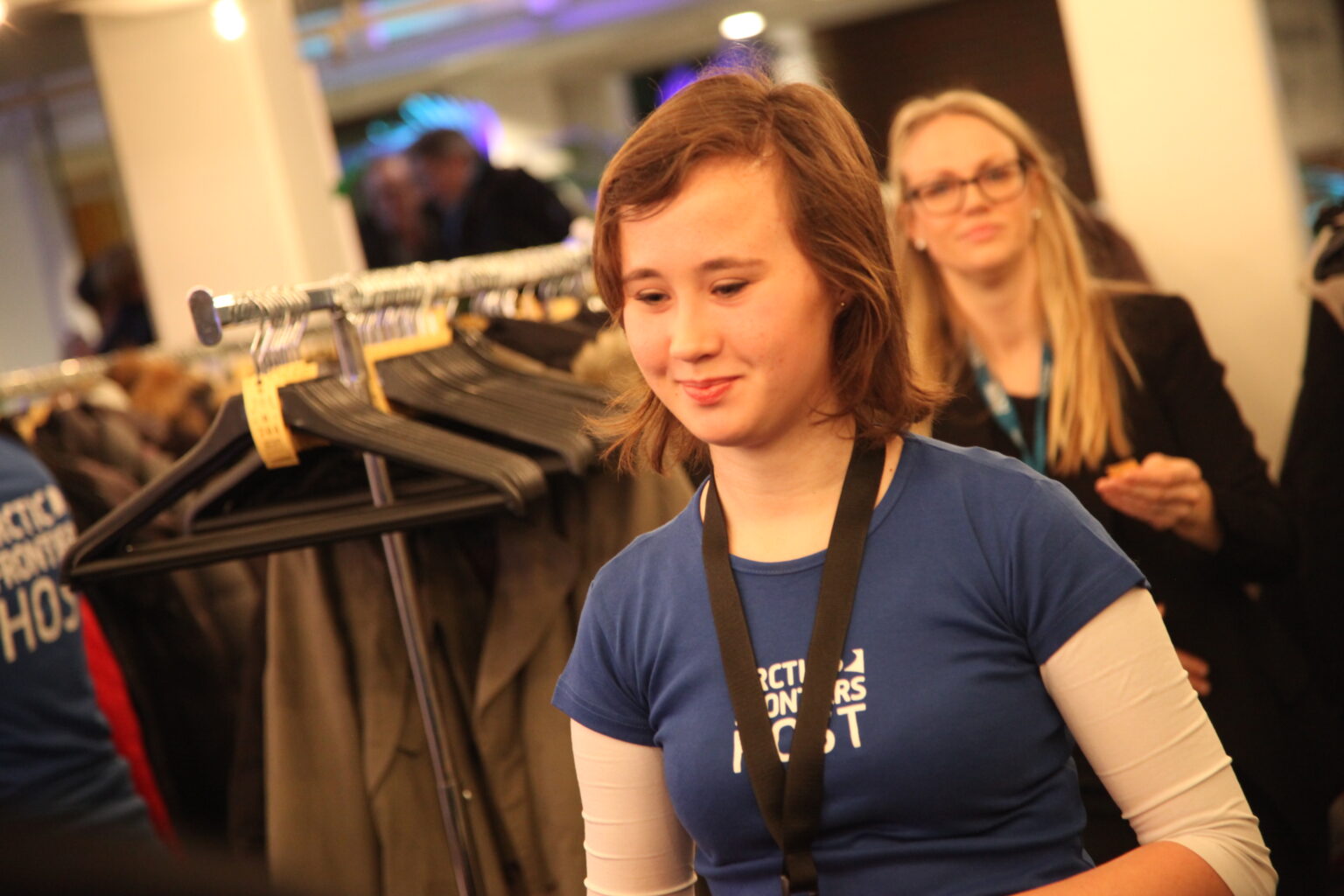 Arctic Frontiers - A young Arctic Frontiers volunteer wearing a blue host t-shirt and a lanyard, with a woman in the background.